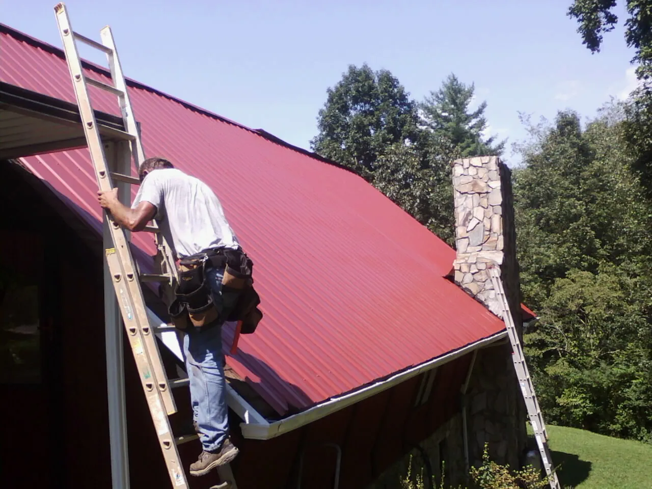 Roofer on a ladder beside a red metal roof and stone chimney — Soffit & Fascia Repair by Roofing Brothers in Luverne